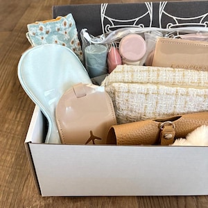 May include: A gift box filled with various items, including a light blue eye mask, a beige passport holder, and a small mirror. Other items include a pair of socks, a small candle, and a cosmetic container. The box is white and the items are arranged inside.