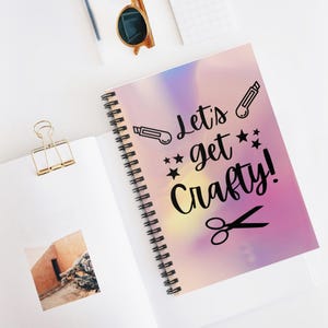 May include: A pink and purple spiral notebook with the text "Let's get crafty!" in black. The notebook has a holographic finish and features a pair of scissors and two craft knives.