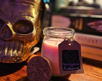 May include: A pink candle in a glass jar with a cork lid. The candle has a brown paper tag with the text "handcrafted by Eatherrie" and a skull graphic.
