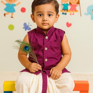 Puede incluir: Un niño pequeño con un atuendo tradicional indio. El conjunto incluye un chaleco sin mangas con botones de color púrpura oscuro, combinado con pantalones dhoti blancos con ribetes púrpuras. El niño sostiene una pluma de pavo real y está sentado en un banco colorido.