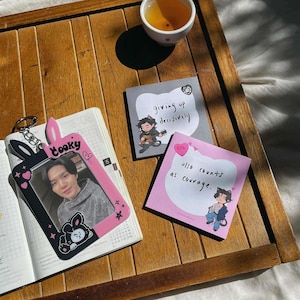 Puede incluir: Una bandeja de madera contiene un portafotos rosa y negro con una foto, un cuaderno y dos notas adhesivas. El portafotos tiene un diseño de orejas de conejo. Las notas adhesivas tienen ilustraciones y texto.