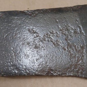 May include: A weathered, dark gray axe head with a rectangular shape and a slightly curved edge. The metal surface has a rough, textured appearance, suggesting age and use. The axe head is likely for woodworking or splitting.