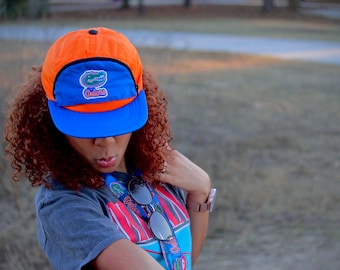 Puede incluir: Una mujer con una gorra de béisbol azul y naranja con un logotipo de caimán y la palabra "Gators" en ella. Lleva una camiseta gris con un estampado gráfico.
