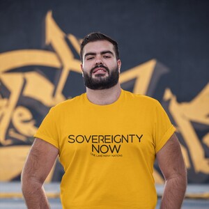 May include: A man wearing a yellow t-shirt with the text "Sovereignty Now" and the words "One Land Many Nations" printed below. He is standing in front of a graffiti wall.