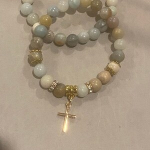 May include: Two beaded bracelets with a gold cross charm. The bracelets are made of light blue, beige, and white beads.