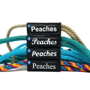 May include: Four black and white labels with the word "Peaches" on them are attached to four different coloured ropes. The ropes are beige, teal, turquoise, and rainbow.