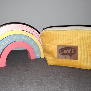 Könnte beinhalten: Ein senfgelbes Cord-Etui mit einem braunen Lederaufnäher "LOVE". Daneben ein Holzregenbogen mit dem deutschen Text "Du machst die Welt bunter."