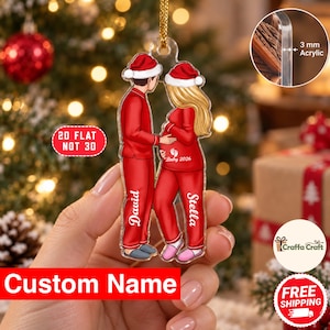 May include: A flat, 3 mm thick acrylic Christmas ornament featuring a cartoon illustration of a couple in red pajamas and Santa hats. The person on the left is labeled Dawid, and the person on the right is labeled Stella with Baby 2026 text.