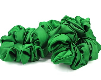 May include: Two vibrant emerald green hair scrunchies made of a smooth, satin-like fabric. Each scrunchie features a gathered, voluminous design with a soft sheen that reflects light across the folds of the fabric, creating a textured appearance.