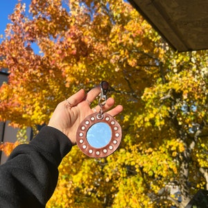 May include: A round, brown leather keychain with a small mirror in the center. The mirror is surrounded by silver studs. The keychain is hanging from a silver clip.