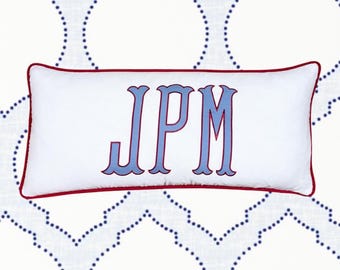May include: White rectangular pillow with red trim and blue embroidered letters "JPM". The pillow is set against a white background with a blue dotted pattern. A decorative accent for a bed or sofa.