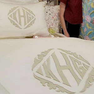 May include: Cream-colored bedding set featuring a monogram design. The set includes a pillow and a decorative emblem with the letters KHG in a stylized font. The emblem is in a light beige color, and the bedding has a subtle striped border.