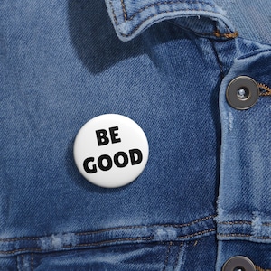 Könnte beinhalten: Ein weißer Ansteckbutton mit den Worten "BE GOOD" in fetten schwarzen Buchstaben. Der Button ist an einer blauen Jeansjacke befestigt. Die Jacke hat sichtbare Nähte und Metallknöpfe.