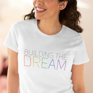 Può includere: Maglietta bianca con il testo "BUILDING THE DREAM" in colori arcobaleno.