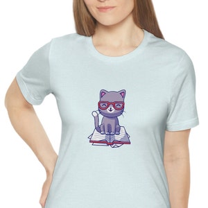 Puede incluir: Una camiseta azul claro con una ilustración de dibujos animados de un gato gris con gafas rojas sentado en un libro.