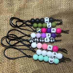 May include: A set of seven colorful silicone bead necklaces with black cords and letter beads spelling out the names "HHJ", "MPE", "H", and "SHS".