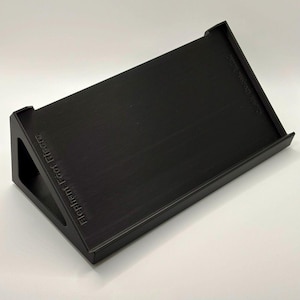 May include: A black, angled stand with the text "Elephant Foot Risers" and "Cute Desk Things" printed on the sides. The stand has a rectangular top with raised edges and a textured surface.