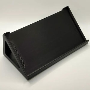 May include: A black, wedge-shaped stand with a textured surface and a raised front edge. The side features the embossed text Elephant Foot Risers, while the upper right corner displays the embossed text Olli Goods LLC.