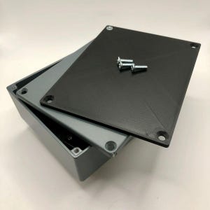 May include: A gray rectangular electrical enclosure with a black lid and two silver screws. The lid is partially open, revealing the interior of the box. The box is designed to protect electrical components.