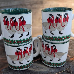 Può includere: Set di quattro tazze in ceramica bianca con bordo verde. Ogni tazza presenta un design festivo con tre oche che indossano fiocchi rossi e un bordo di agrifoglio.