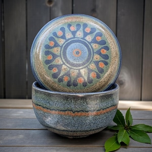 May include: A ceramic bowl and lid set with a blue, green, and orange floral design. The bowl has a textured surface with an orange stripe. The lid features a detailed circular pattern. The set is on a wooden surface.