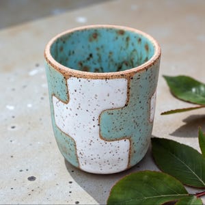 Puede incluir: Una taza de cerámica hecha a mano con un diseño único. La taza presenta un exterior turquesa con un patrón geométrico blanco. El borde tiene un acabado natural sin esmaltar. Ideal para café, té u otras bebidas.