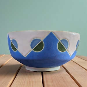May include: A ceramic bowl with a geometric design. The bowl features a white rim, a blue base, and a pattern of blue and green circles and triangles. The bowl is sitting on a wooden surface.