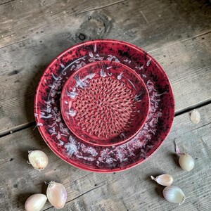 May include: A red ceramic garlic grater set with a matching plate. The grater has a textured surface for efficient grating. The set is red with black and white accents. Several garlic cloves are scattered around the set on a wooden surface.