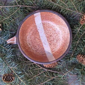 May include: A brown ceramic bowl with a white stripe down the center. The bowl has a speckled glaze and a slightly rough texture. The bowl is sitting on a bed of pine needles.
