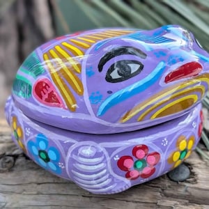 Könnte beinhalten: Eine violette Keramik-Frosch-Schmuckschatulle mit farbenfrohen Maldetails. Der Frosch hat einen geschlossenen Mund und große Augen. Die Schachtel ist mit Blumen und Spiralen verziert.