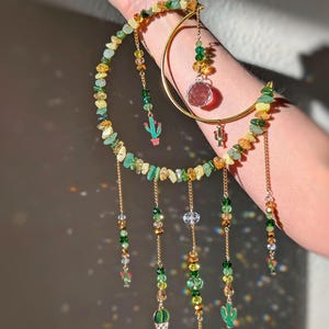 May include: A decorative suncatcher featuring a gold crescent moon and a smaller gold hoop. The suncatcher is adorned with green, yellow, and clear beads, along with small cactus charms. Strands of beads hang from the moon and hoop.