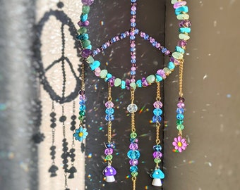 Pode incluir: Um apanhador de sol em forma de sinal de paz com um prisma de cristal pendurado na parte inferior. As contas são em tons de turquesa, roxo e verde. Também há pequenos amuletos de flores e cogumelos pendurados na parte inferior.