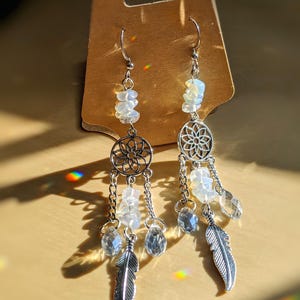 May include: A pair of silver-toned dangle earrings. Each earring features a dreamcatcher design, clear crystal teardrops, and a silver feather charm. The earrings also have white, irregular-shaped stones and are displayed on a brown card.