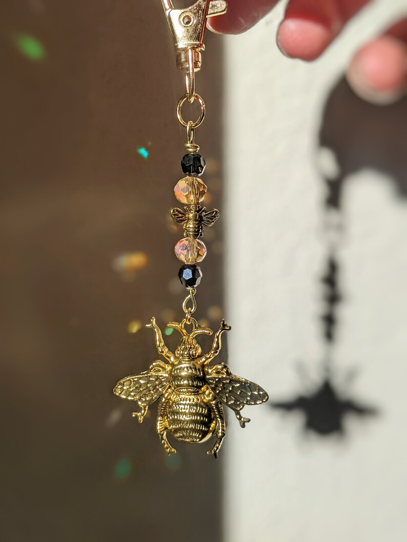 May include: A gold-colored bee charm keychain with a lobster clasp. The charm features a large, detailed bee pendant, with smaller bee and bead accents in black and amber. The keychain is suspended from a gold-tone metal chain.