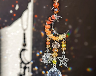May include: A silver crescent moon charm with orange, yellow, and clear crystal beads hangs from a silver chain. The charm has a crystal sun and star hanging from it. A clear crystal prism hangs from the bottom of the charm.