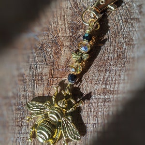 May include: A gold-colored bee charm with a lobster clasp, adorned with amber and black beads. The bee charm is detailed with wings and body segments. This accessory can be used as a keychain or a bag charm.