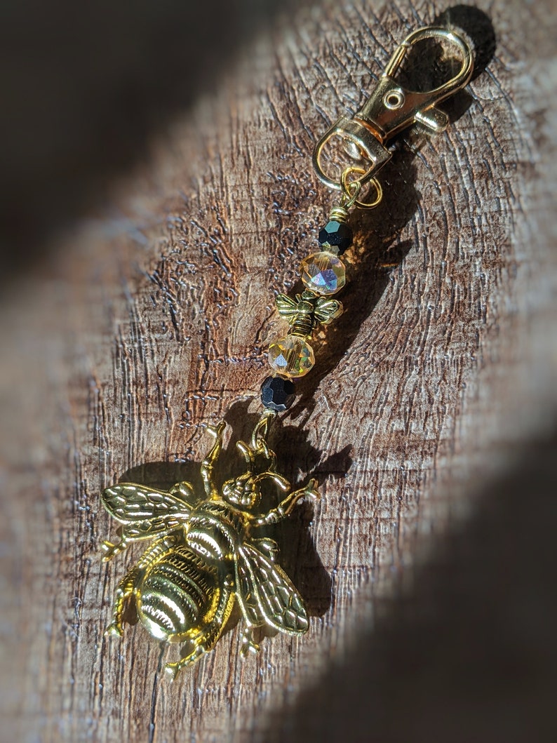 May include: A gold-colored bee charm with a lobster clasp, adorned with amber and black beads. The bee charm is detailed with wings and body segments. This accessory can be used as a keychain or a bag charm.
