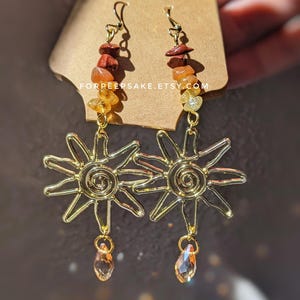 May include: Gold-tone sun-shaped earrings featuring a spiral center and dangling teardrop crystals. The upper portion includes stacked red, orange, and yellow chip beads. The text FORPEEPSAKE.ETSY.COM is printed on the brown display card.