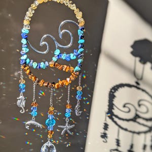 May include: A crystal suncatcher with a silver wave design and beads in shades of blue, orange, and yellow. The suncatcher has a crystal prism hanging from the bottom.