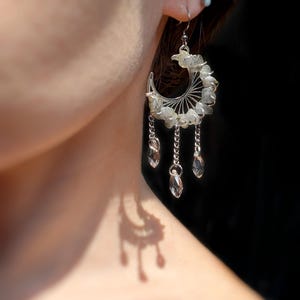 May include: A silver-toned crescent moon earring featuring a wire-wrapped frame adorned with small, pale green stone chips. Three delicate silver chains hang from the bottom of the crescent, each ending in a clear, faceted, teardrop-shaped crystal bead.