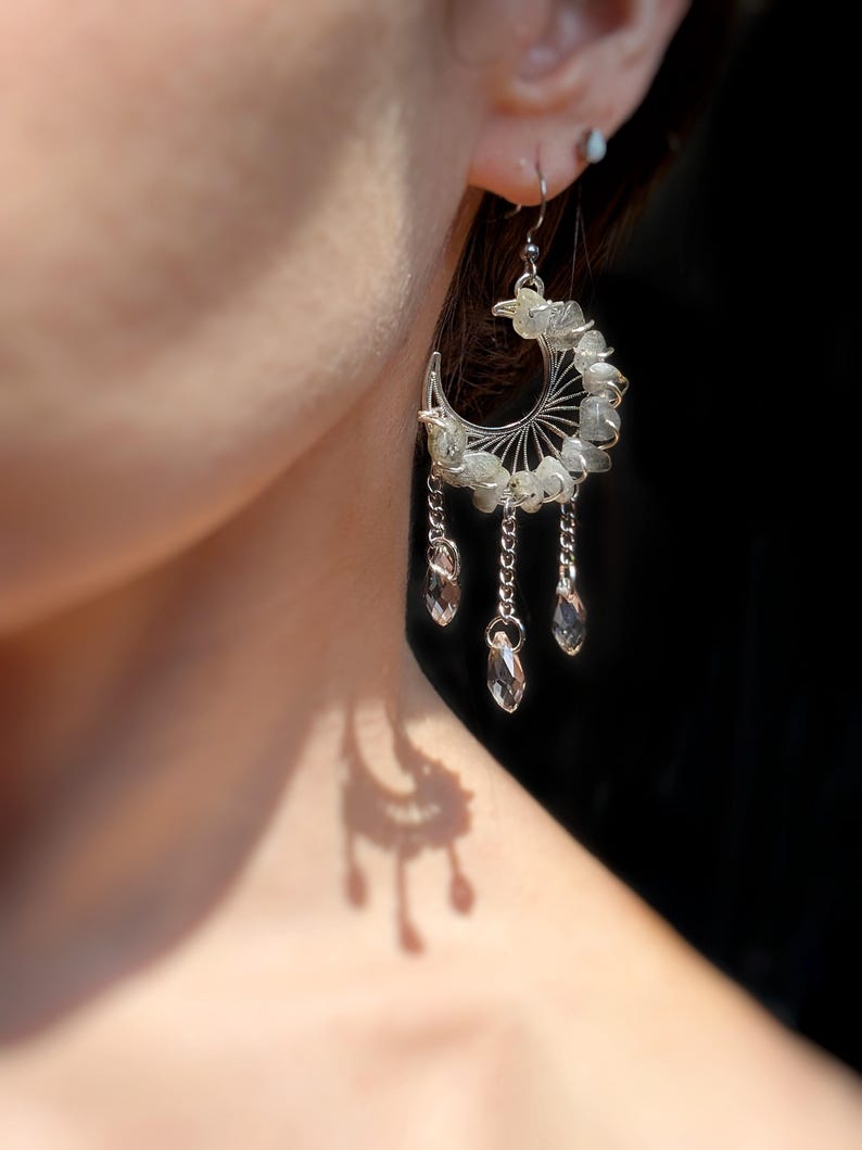 May include: A silver-toned crescent moon earring featuring a wire-wrapped frame adorned with small, pale green stone chips. Three delicate silver chains hang from the bottom of the crescent, each ending in a clear, faceted, teardrop-shaped crystal bead.