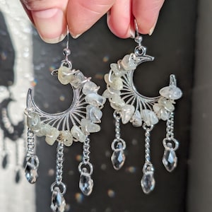 May include: A pair of silver-toned crescent moon earrings featuring wire-wrapped pale green tumbled stones along the curve. Each earring includes three dangling silver chains ending in clear, faceted teardrop-shaped crystal beads.