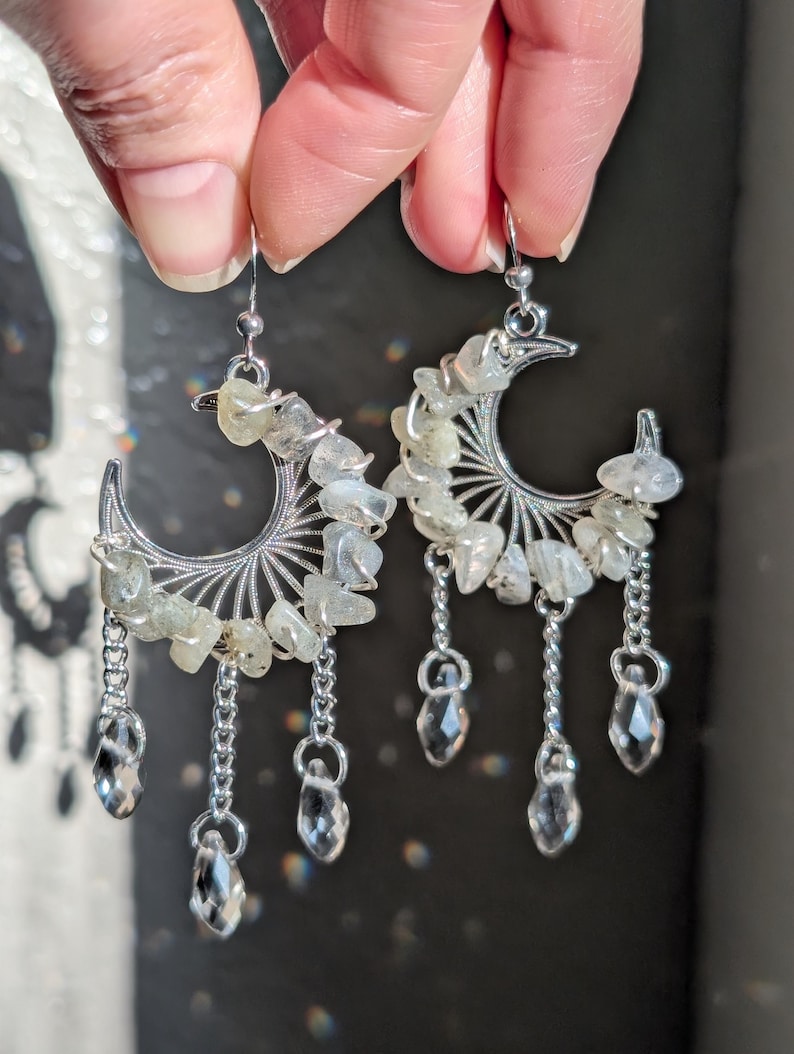 May include: A pair of silver-toned crescent moon earrings featuring wire-wrapped pale green tumbled stones along the curve. Each earring includes three dangling silver chains ending in clear, faceted teardrop-shaped crystal beads.