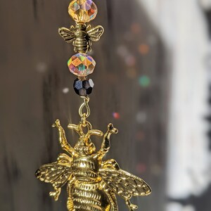 May include: A gold-colored bee charm with a detailed design, hanging from a chain with black and iridescent crystal beads. The charm is a decorative accessory, possibly for a necklace or keychain.
