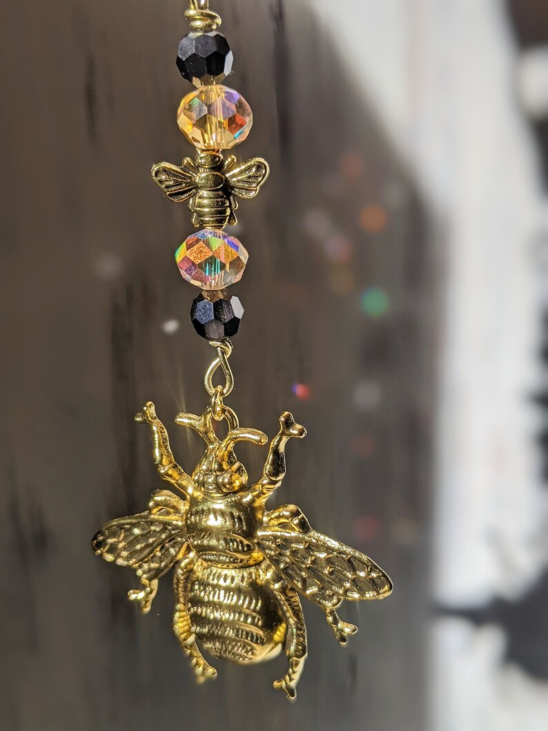 May include: A gold-colored bee charm with a detailed design, hanging from a chain with black and iridescent crystal beads. The charm is a decorative accessory, possibly for a necklace or keychain.