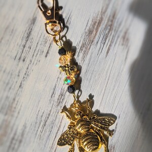 May include: A gold-colored bee charm with a lobster clasp, adorned with small beads and a smaller bee charm. The large bee charm has detailed wings and body segments. The background is a white, weathered wood surface.