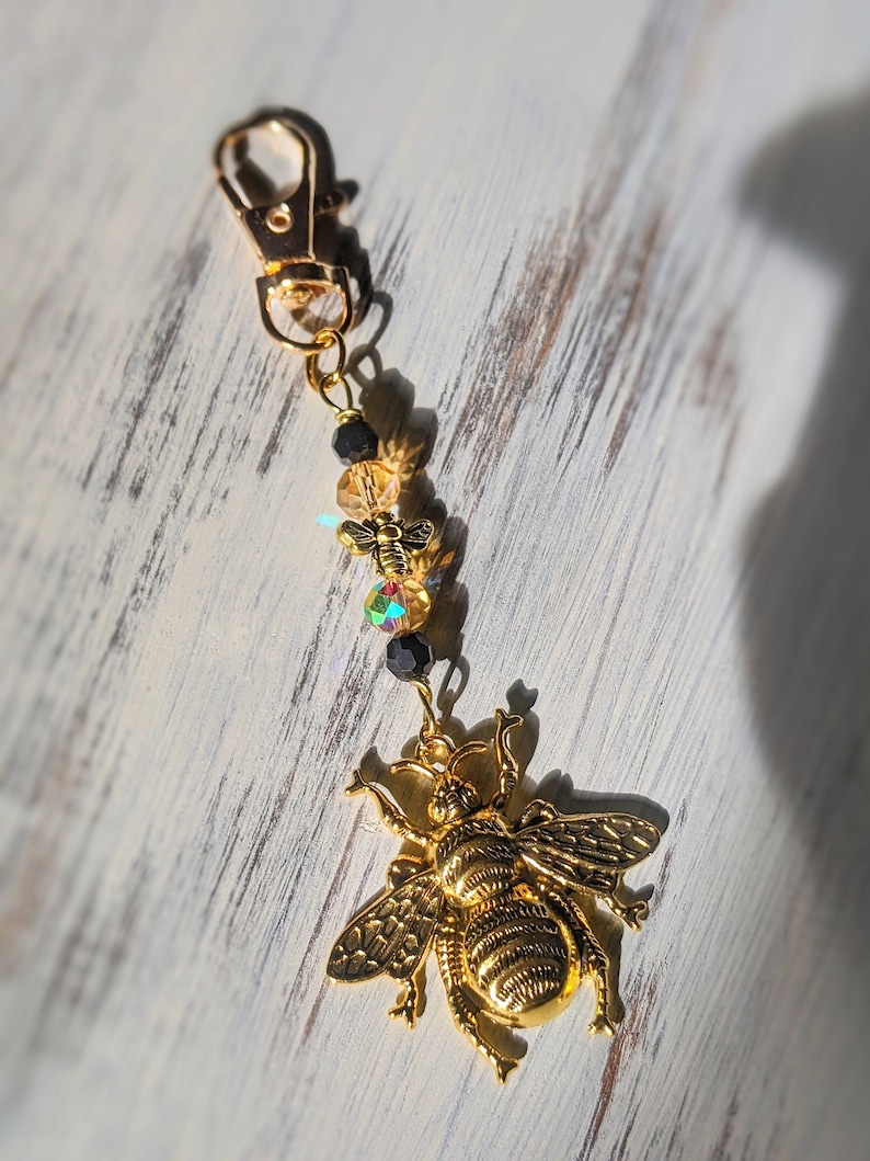 May include: A gold-colored bee charm with a lobster clasp, adorned with small beads and a smaller bee charm. The large bee charm has detailed wings and body segments. The background is a white, weathered wood surface.
