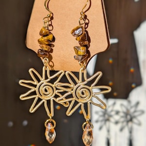 May include: Gold-colored wire earrings featuring a sunburst design with a spiral center and dangling teardrop-shaped crystals. The earrings also have a strand of brown and gold-colored stones. The earrings are displayed on a brown card.