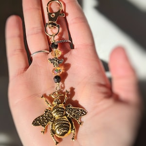 May include: A gold-colored bee charm keychain with a lobster clasp. The keychain features a large, detailed bee pendant, iridescent and black beads, and a gold-tone clasp. The bee pendant is the focal point, showcasing intricate wing and body details.