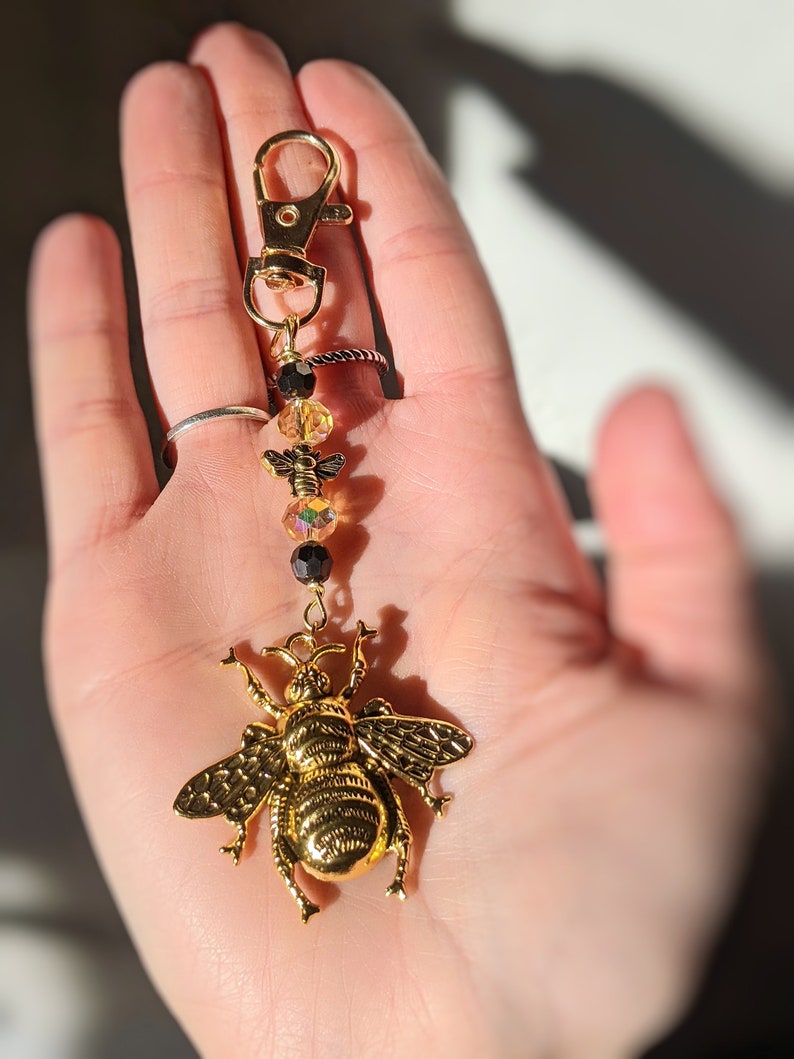 May include: A gold-colored bee charm keychain with a lobster clasp. The keychain features a large, detailed bee pendant, iridescent and black beads, and a gold-tone clasp. The bee pendant is the focal point, showcasing intricate wing and body details.